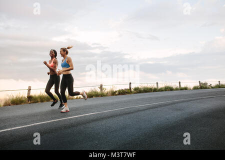 Zwei Athleten laufen früh am Morgen auf der Straße mit bewölktem Himmel im Hintergrund. Fitness-Frauen, die auf der Straße joggen. Stockfoto