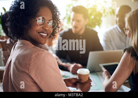 Lächelnd afrikanischen Geschäftsfrau Blick über ihre Schulter, während bei einer Tabelle mit Kollegen im Hintergrund sitzen Stockfoto
