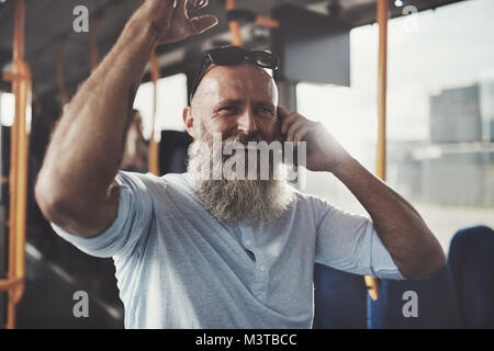 Reifer Mann mit langem Bart Lachen beim Stehen auf einem Bus über sein Handy während seines morgendlichen Fahrt Stockfoto
