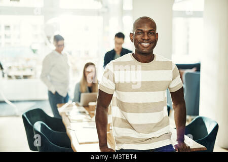 Junge afrikanische Designer selbstbewusst lächelnd während stützte sich auf eine Tabelle in einem Büro mit Kollegen im Hintergrund arbeiten Stockfoto