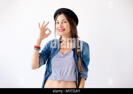 Lächelnd fröhliches Mädchen, mit geflochtenen langen kastanienbraunen Haar zu tragen Jeanshemd, Oberteil und Hut, abs, okay Zeichen auf weißem Hintergrund in Stud Stockfoto