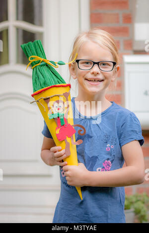 Ein blondes Mädchen in Gläsern freut sich auf seinen ersten Tag an der Schule und wirft mit der Schultasche vor der Haustür. Stockfoto