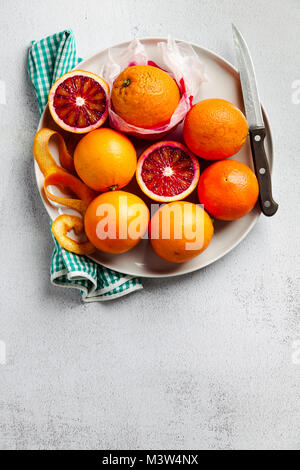 Eine Gruppe von Orangen auf einem Teller. Einige von ihnen geschnitten. Messer und Handtuch. Auf einem Stein Hintergrund. Eine gesunde Sommer Tabelle, die Vorbereitung von frisch gepressten j Stockfoto