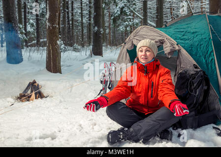Junge aktive Tourist auf einer Kampagne sitzt in der Nähe von einem Zelt im Winter Forest Stockfoto