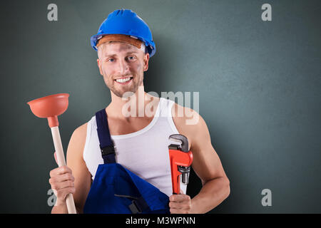 Portrait Of Happy Klempner Holding Schraubenschlüssel und Kolben auf grauem Hintergrund Stockfoto