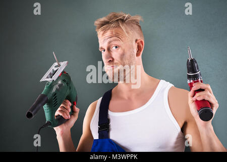 Porträt einer männlichen Techniker Holding elektrische Bohrmaschine und Schraubendreher auf grauem Hintergrund Stockfoto