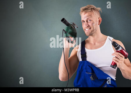 Porträt einer männlichen Techniker Holding elektrische Bohrmaschine und Schraubendreher auf grauem Hintergrund Stockfoto