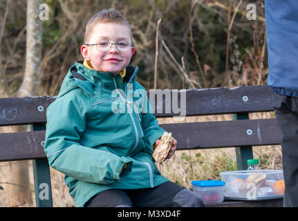 6 Jahr alten lächelnde junge Brille sitzen auf einer Bank bei einem Picknick Mittagessen. Er hält einen Schinken und Käse rollen, im Winter, Schottland, UK Stockfoto