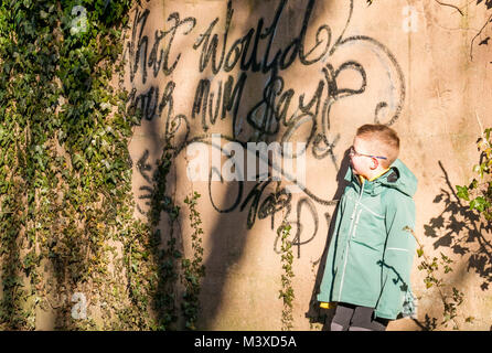 6 Jahre alter Junge mit Brille neben Sprühfarbe Graffiti an der Wand, das liest Was Wäre deine Mama sagen?, Schottland, Großbritannien Stockfoto