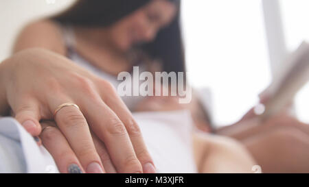 Trauringe auf Händen eines glückliches junges attraktives Paar auf der Couch, das Mädchen, das ein Buch, das auf ihrem Schoß liegt liest Stockfoto