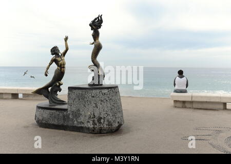 Eine einsame Frau sitzt auf dem Puerto Vallarta malecon mit Möwen am Meer in der Nähe fliegen und eine Statue von Triton merman und Nereida mermaid hinter ihr. Stockfoto