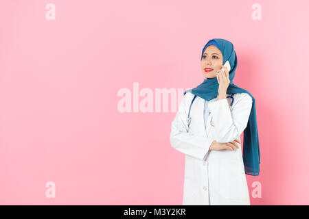 Lächelnd ziemlich muslimische Frau Doktor über Mobile Smartphone Aufruf für Kollegen diskutieren Krankenhaus arbeiten, wenn Sie in rosa Hintergrund stehen. Stockfoto