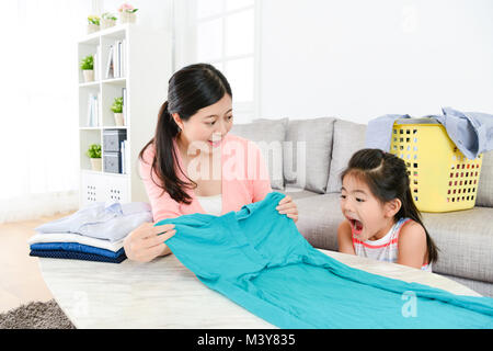 Elegante Schönheit Hausfrau falten Familie Kleidung in Wohnzimmer und ihre kleine Tochter Gefühl überrascht. Stockfoto