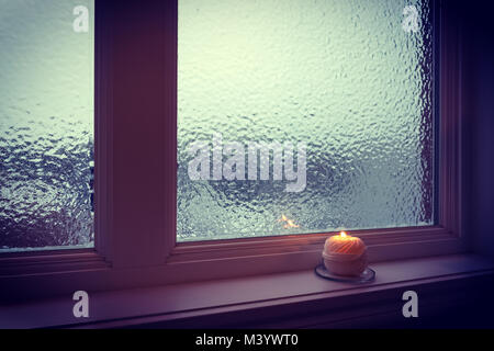 Kerze in der Nähe von einem Fenster aus Milchglas in der Dämmerung. Winter Abend. Stockfoto