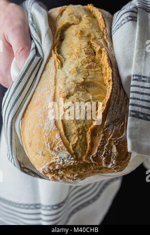 Männliche Baker Holding in den Händen frisch gebackene Handgefertigte Rustikales Brot Brot verpackt in weißer Bettwäsche Handtuch. Natürliches Licht authentischen Stil. Lifestyle Foto. Stockfoto