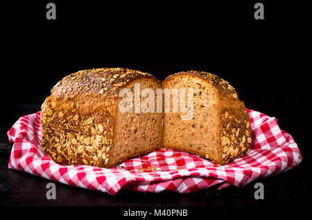 Zwei Hälften Getreidespeicher Dinkel Brot auf roten und weißen Handtuch für die Küche und schwarzem Holz- Hintergrund. Vorderansicht, Nahaufnahme. Stockfoto