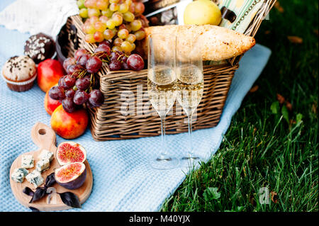 Picknick im Park auf einer Wiese, leckeres Essen: Korb, Wein, Trauben, Pfirsiche, Baguette, Cupcakes, Feigen, Käse, Blaue Tischdecke, Weinglas mit Champa Stockfoto