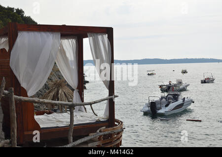 Cabana Bett mit Baldachin und weißen Vorhängen mit Blick auf einen See mit einem Schnellboot im Sommer Stockfoto