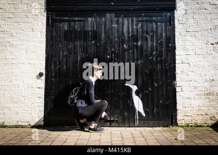 Hipster Frau mit schwarzen und Sonnenbrille auf der Suche nach einem kleinen weißen Vogel Zeichnung (ähnlich wie ein Reiher) auf einem großen schwarzen Holz Garagentor Stockfoto