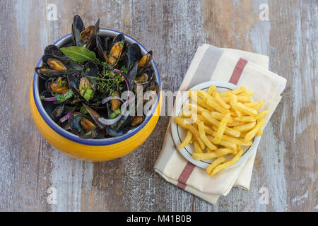 Französische Miesmuschel mit Kräutern in einem gelben Schüssel mit Pommes frites ramekin Glas auf Serviette Fisch und Meeresfrüchte in einer Holzplatte Hintergrund Stockfoto