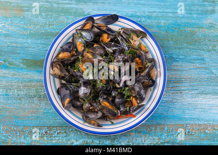 Gedämpfte Muscheln in Weißwein mit Kräutern, Fisch und Meeresfrüchte. In weißer Teller serviert, bis auf Blau Holz- Hintergrund Stockfoto