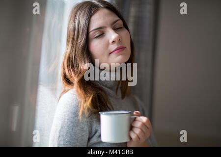Junge erwachsene Frau lehnte sich auf Fenster mit Becher in der Hand Stockfoto