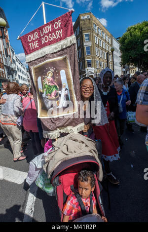 Eine Frau nimmt teil an der Pfarrei die Prozession am Fest Unserer Lieben Frau vom Berge Karmel in der Italienischen Kirche in Clerkenwell mit einem Kinderwagen und einem Banner der Gottesmutter vom Rosenkranz. Stockfoto