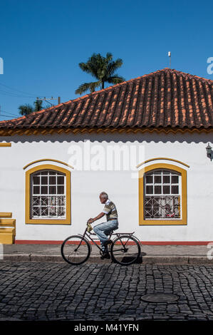 Ein alter Mann, eine Steel Bike auf einer Straße mit Kopfsteinpflaster vor einem Haus im Kolonialstil in Antonina, Brasilien Stockfoto