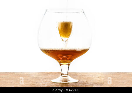 Bier ist in ein Glas gießen, der in eine andere sehr große Glas, Erstellen von Schaum und Blasen auf dem hölzernen Textur an der Unterseite und weißem Hintergrund Stockfoto