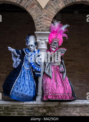 Zwei Frauen neben einer Spalte, halten die Fans, und das Tragen von Hand bemalten Masken und kunstvollen blau und rosa Kostüme im Karneval von Venedig Stockfoto