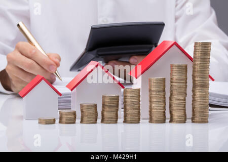 Nahaufnahme der Geschäftsmann Berechnung Rechnung mit verschiedenen Größe Häuser und Stapel von Münzen auf Tisch Stockfoto
