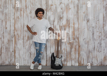 Kreative Person. Mulatte mann Musiker, mit lockigem Haar steht in der Nähe der Bass Gitarre, gegen retro Holzwand Stockfoto