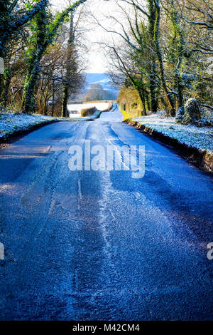 Einer von Bäumen gesäumten Country Lane mit Bergab, aspalt Patches auf der Straße, eine Schicht aus weißem Schnee auf jeder Seite auf dem Boden und Bäumen, niedrige Winte Stockfoto