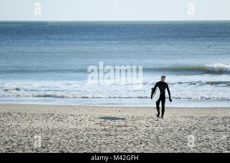 Ein Surfer tragen eine Ganzkörper Anzug, Spaziergänge am Strand, vom Meer, mit seinem Surfbrett auf einem kalten helle Winter Tag in Bournemouth, Großbritannien Stockfoto