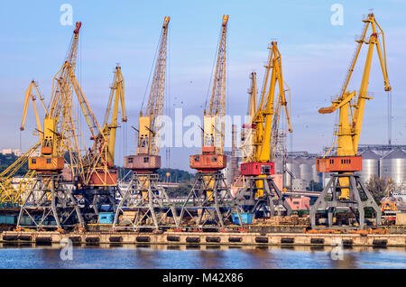 Gelben Krane, die in einem Seehafen vor dem Hintergrund von Metall Getreidespeicher und ein blauer Himmel mit Wolken und fliegen Scharen von Vögeln Stockfoto