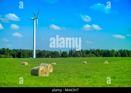 Szene Sommer wind turbine in der Mitte der Wiese voller Strohballen, neben Wald, während der schönen Sonnenuntergang auf Land. Ländliche Landschaft, wi Stockfoto