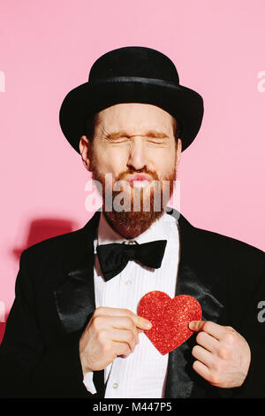 Lustiger Kerl in Tux und Hut schmollen, ein Kuss und hält ein rotes Herz Stockfoto