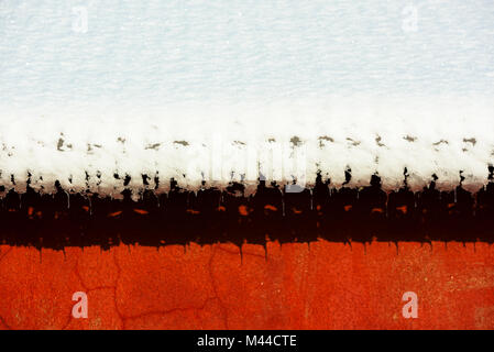 Schnee und Eiszapfen langsam gleiten von einem Dach an einem warmen Tag. Rot bunte Wand hinter Schnee. Stockfoto