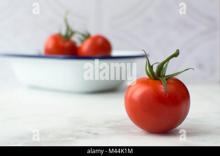 Leuchtend rote Tomaten in eine Schüssel mit weißen Negative füllen. Rohstoff, helles Rot mit Blau und Weiß Emaille Teller in auf einem weißen Marmor Oberfläche geschossen. Stockfoto