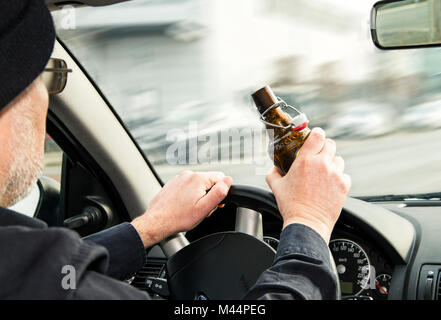 Mann hält eine geöffnete Flasche Bier in der einen Hand und deckt das Lenkrad eines Autos mit dem Anderen. Stockfoto