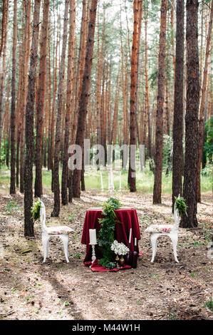 Jäten Dekor. Gedeckter Tisch für zwei serviert mit zwei weißen geschmiedet Stühle im Pinienwald. Dekoration mit Marsala Stoff, Kerzen, Kerzenhalter, Wein Stockfoto