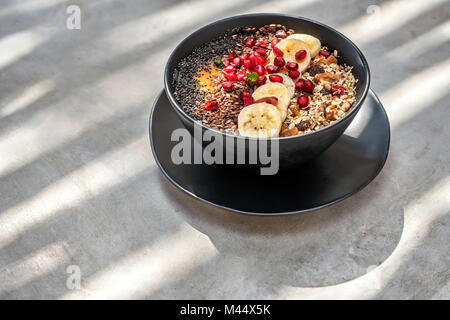 Exotische Lebensmittel Mix in einem schwarzen Schüssel auf den dunklen Teller auf dem Tisch. In der Schüssel Es gibt Scheiben von Bananen, Nüssen und Samen, Granat, Minze und Fruchtfleisch Stockfoto
