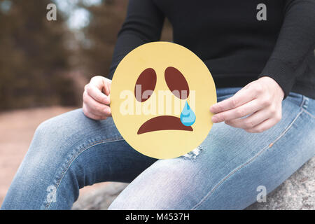 Traurige emoticon Papier in der Hand. Deprimiert Frau mit gedruckten crying Smiley und sitzen auf einem Felsen. Moderne Kommunikations- und Smiley. Stockfoto