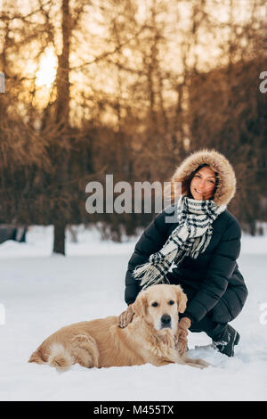 Fotos von lächelnden Mädchen auf Spaziergang mit Hund auf dem Hintergrund der Bäume im Winter Park am Nachmittag Stockfoto