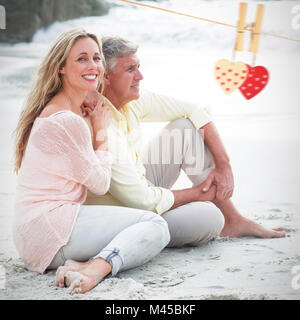 Zusammengesetztes Bild des glücklichen Paares sitzen auf dem sand Stockfoto