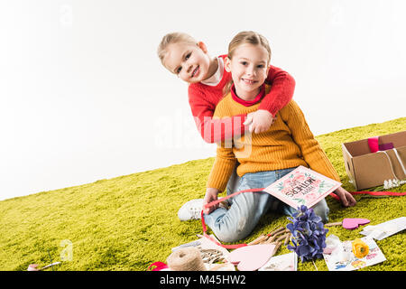 Kleinen Schwestern sitzen auf dem Boden, während Sie Muttertag Grußkarte und umarmt von Weiß isoliert Stockfoto