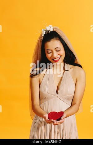Asiatische Braut in beige Wedding Dress hält ein rotes Herz isoliert auf Orange Stockfoto