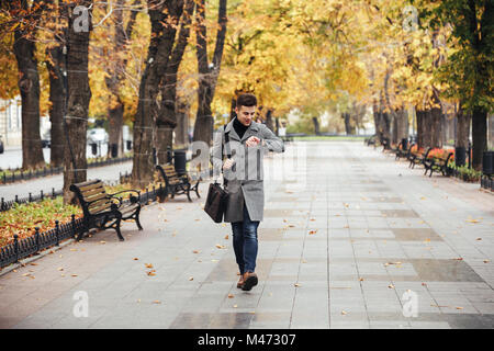 Bild von gut aussehenden Kaukasier Mann im Mantel mit Tasche Bummeln in der City Park und schaut auf seine Uhr Stockfoto