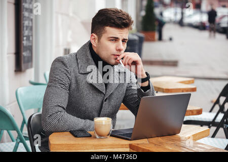 Gut aussehende kaukasischen Typ mit Brüten Sie im Cafe draußen rauchen Zigarette und trinken Cappuccino sitzen Stockfoto