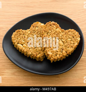 Zwei selbstgebackene herzförmige Erdnuss und Hafer flapjack Kekse für Valentines Tag auf ovalen schwarzen Teller gemacht auf hölzernen Tisch. Stockfoto